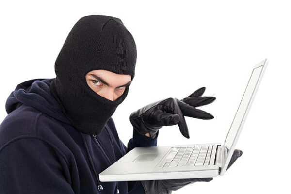Hacker in balaclava standing and typing on laptop on white background